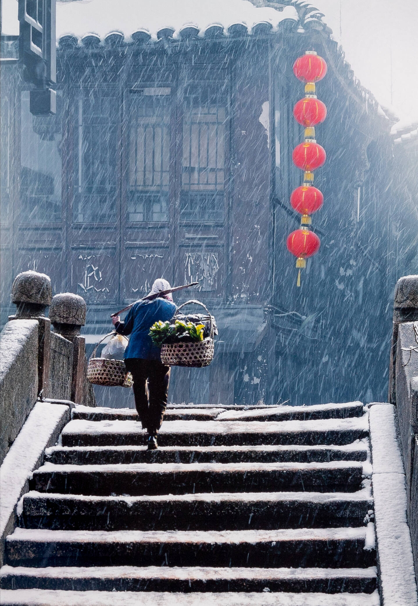 Color photograph of a man carrying to baskets on his shoulders walking up a set of stairs in the snow.